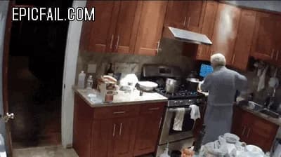 Smoke pours off a pan on the gas stove while an older woman moves around the kitchen.