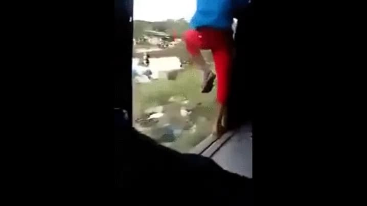 A man hangs out of the open door of a moving train, gripping the handrail as he swings outward.